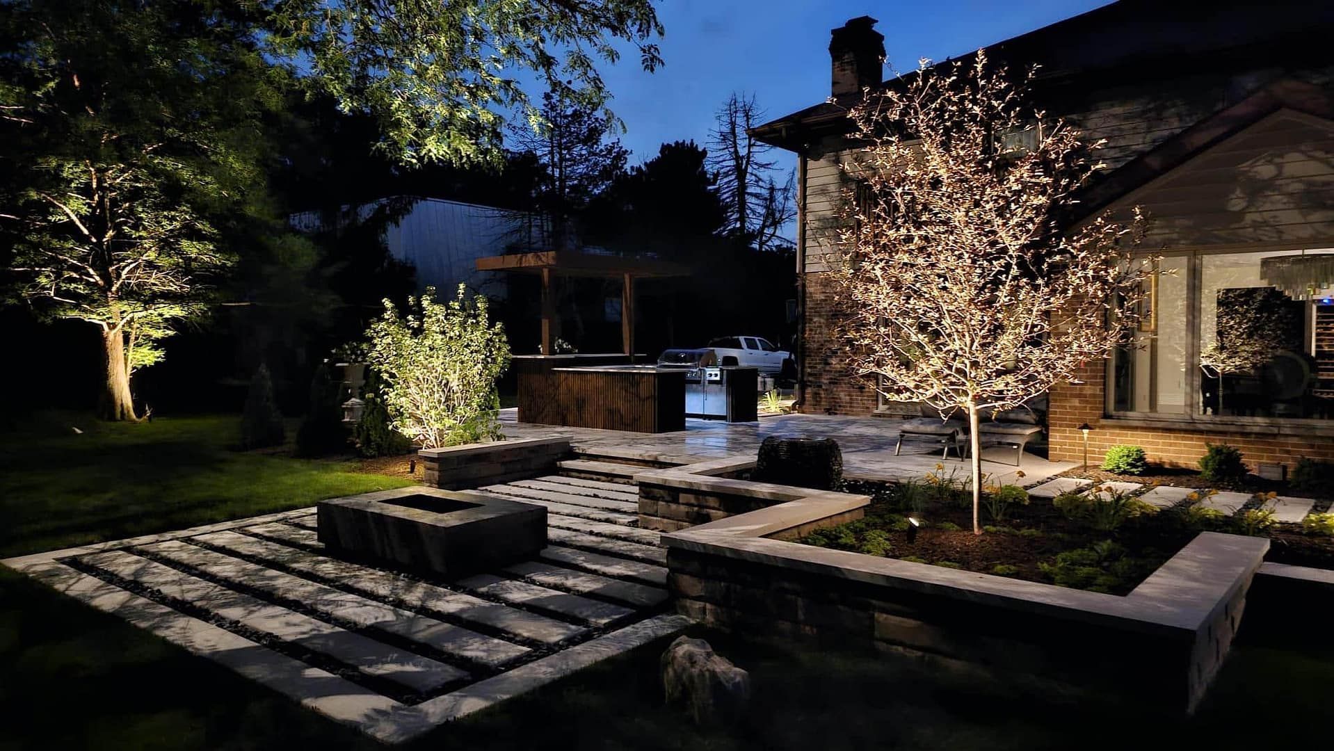 Nighttime backyard patio with landscape lighting illuminating trees, plants, and a stone fire pit.