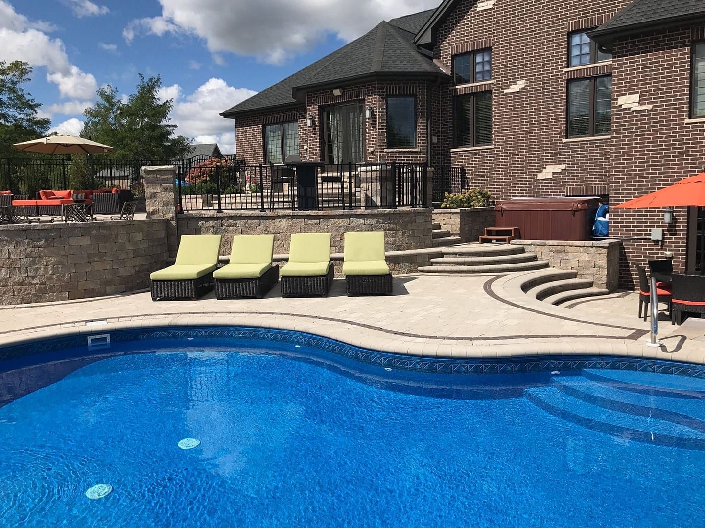 Swimming pool in a backyard with lounge chairs, hot tub, and brick house under a sunny sky.