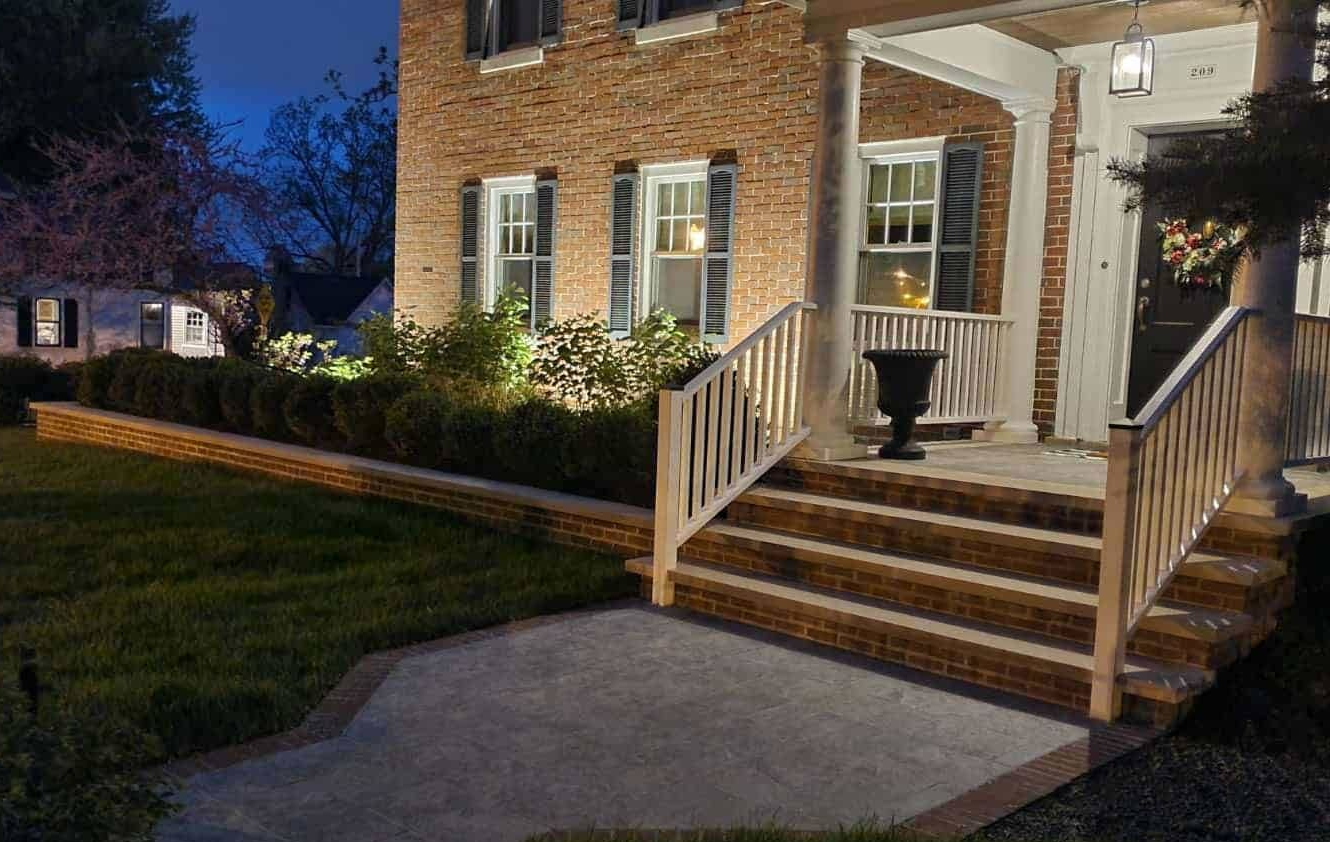 Brick home at night with lit porch and landscaped yard. Concrete steps, shrubs, and a retaining wall.