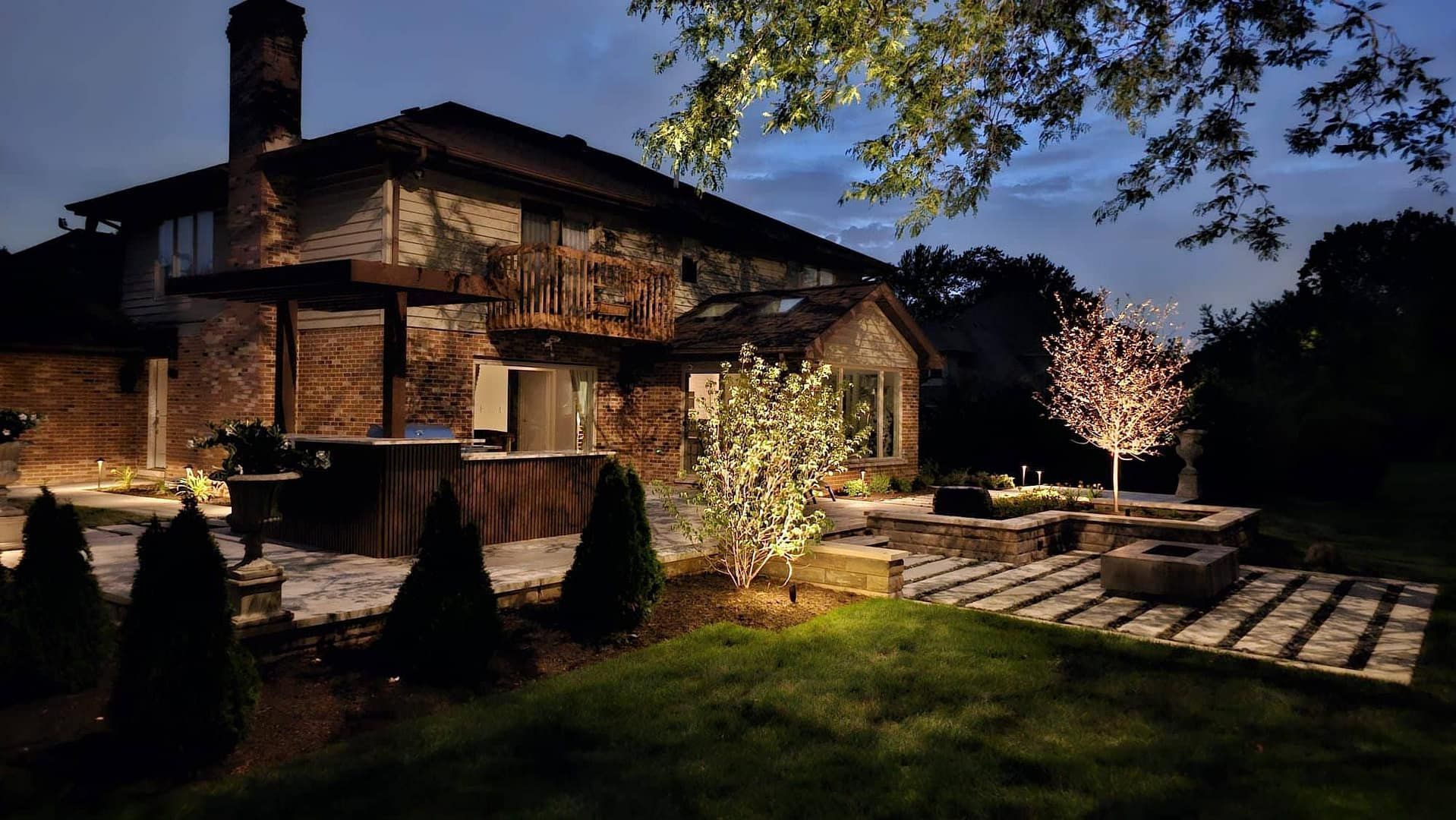 Night view of a brick house with outdoor lighting. A patio and landscaping are illuminated in a dusk setting.