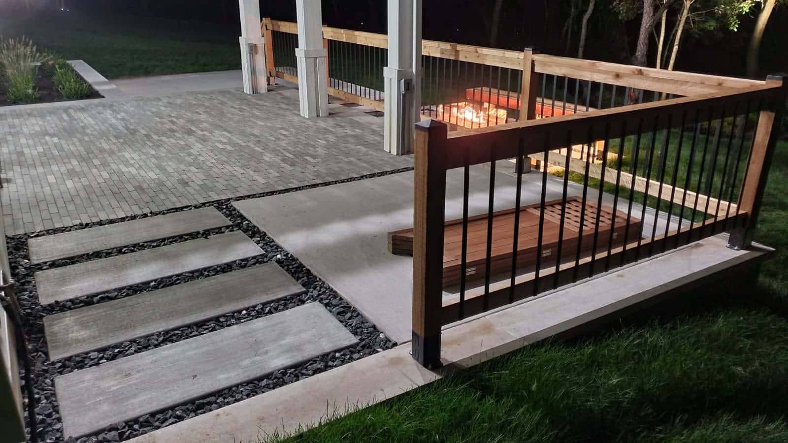 Patio with fire pit and wooden railing at night. Concrete steps with dark pebbles border the walkway.