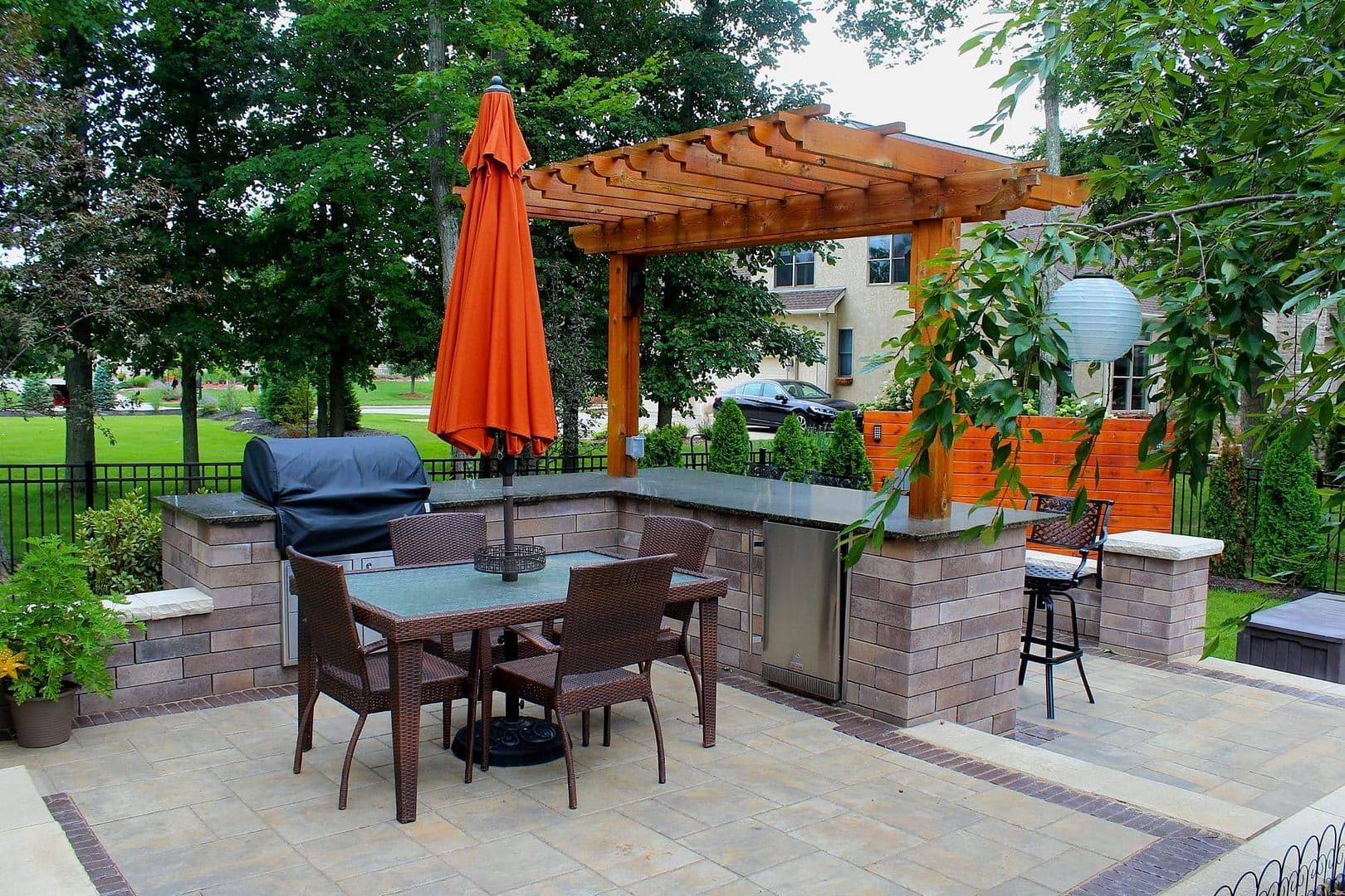 Outdoor patio with grill, table, chairs, orange umbrella, and wooden pergola.
