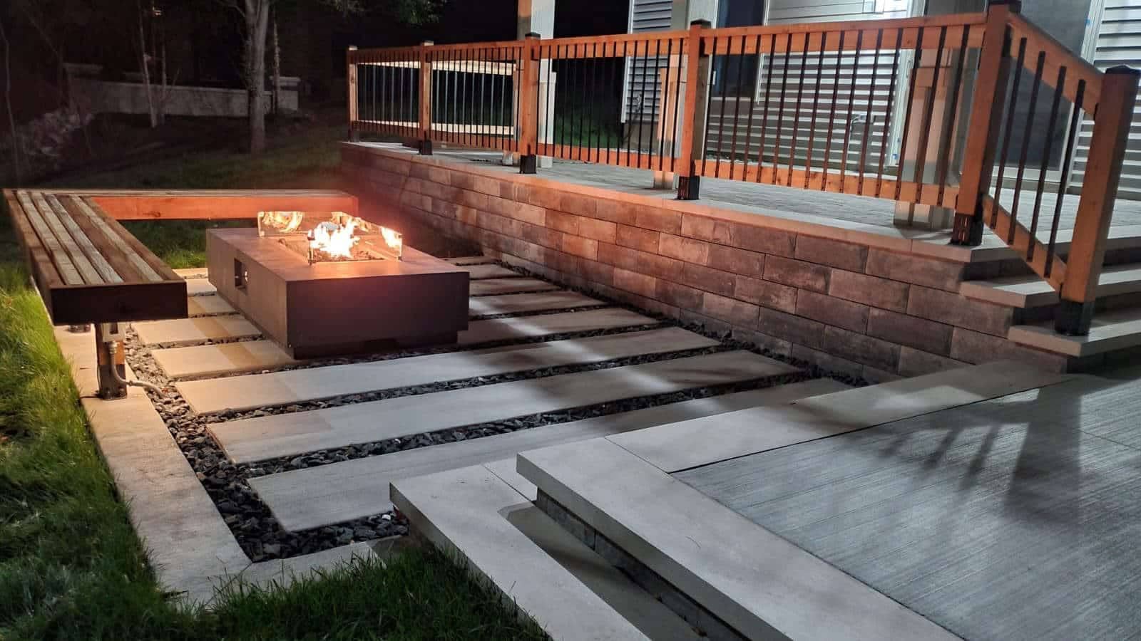 Outdoor fire pit area with concrete pavers, wooden bench, and elevated deck with railings. Nighttime scene.