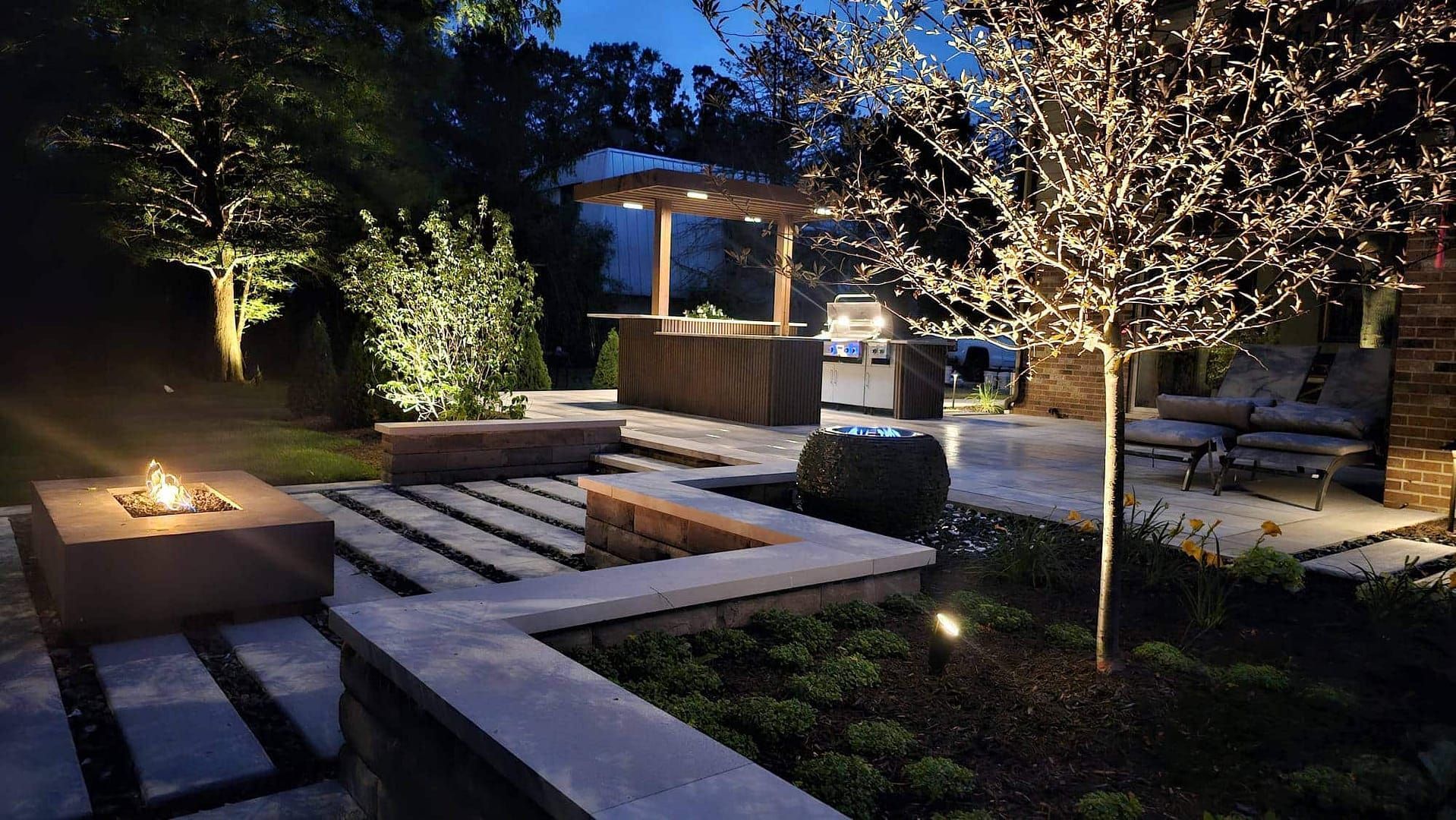 Nighttime backyard scene with fire pit, stone patio, pergola, and illuminated trees.