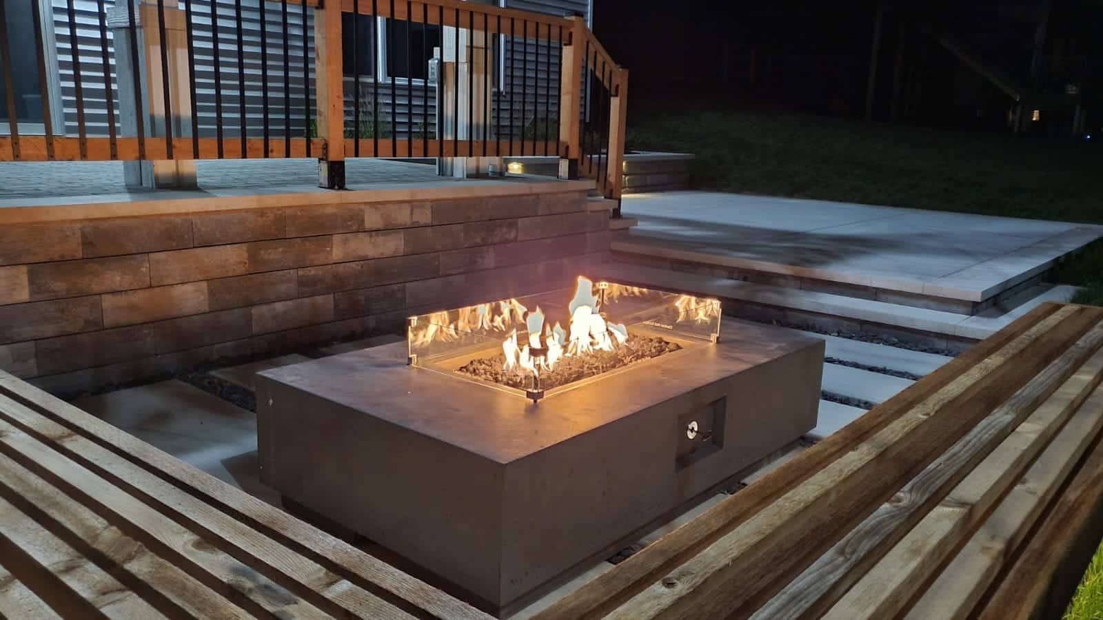 Fire pit with flames surrounded by wooden benches and a tiered patio at night.
