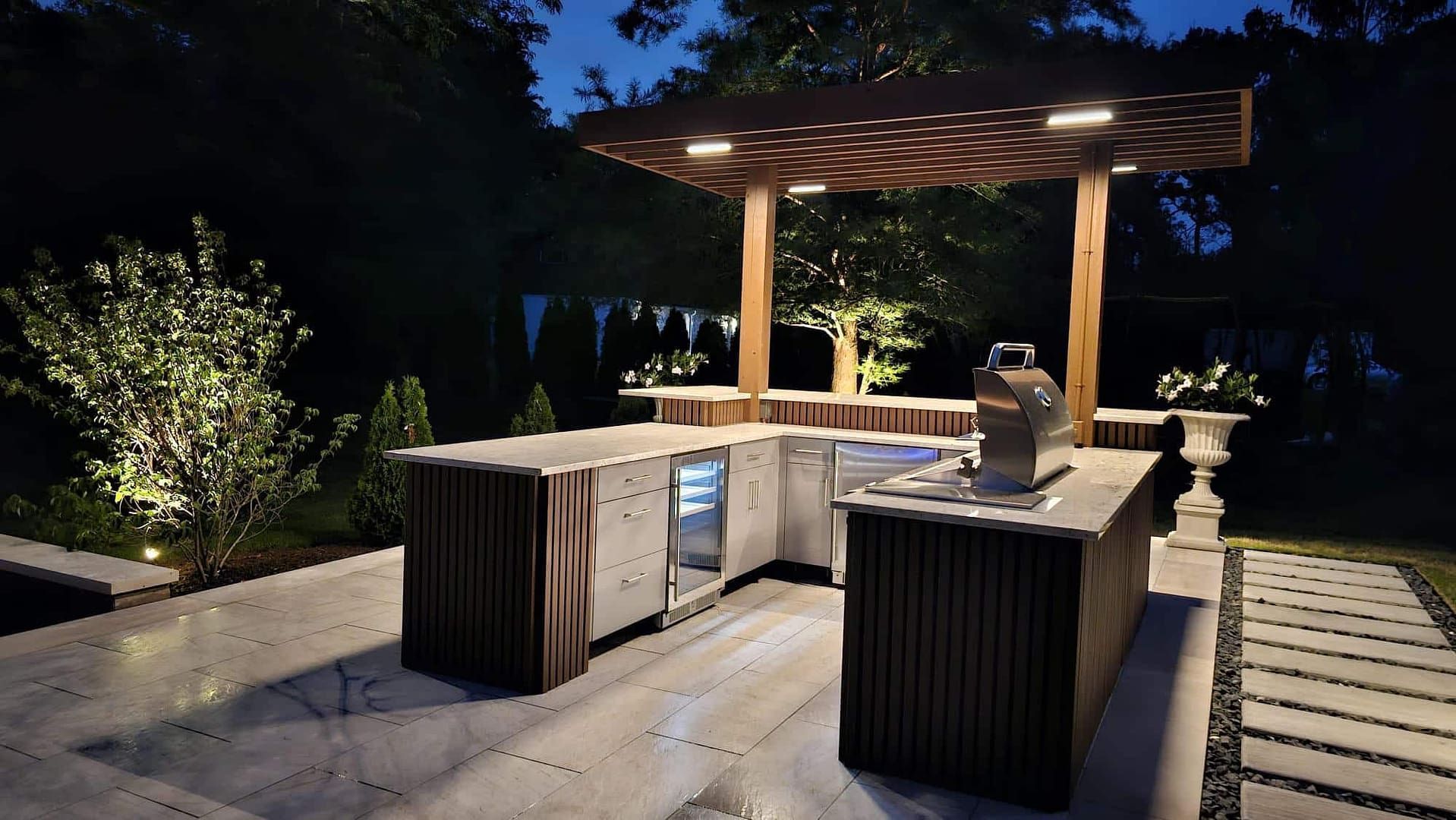 Outdoor bar with overhead lighting at night. Features include grill, refrigerator, and a pergola.