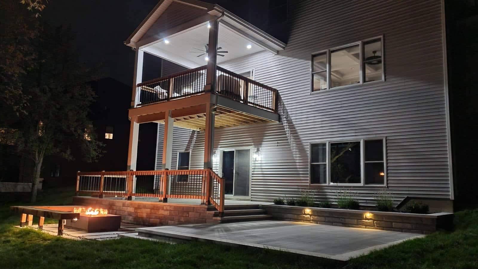 Nighttime view of a two-story house with a lighted deck and patio, fire pit, and backyard.