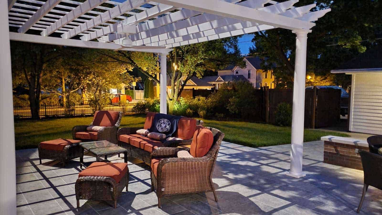 Patio with wicker furniture under a white pergola, lit at dusk; lawn and houses in background.