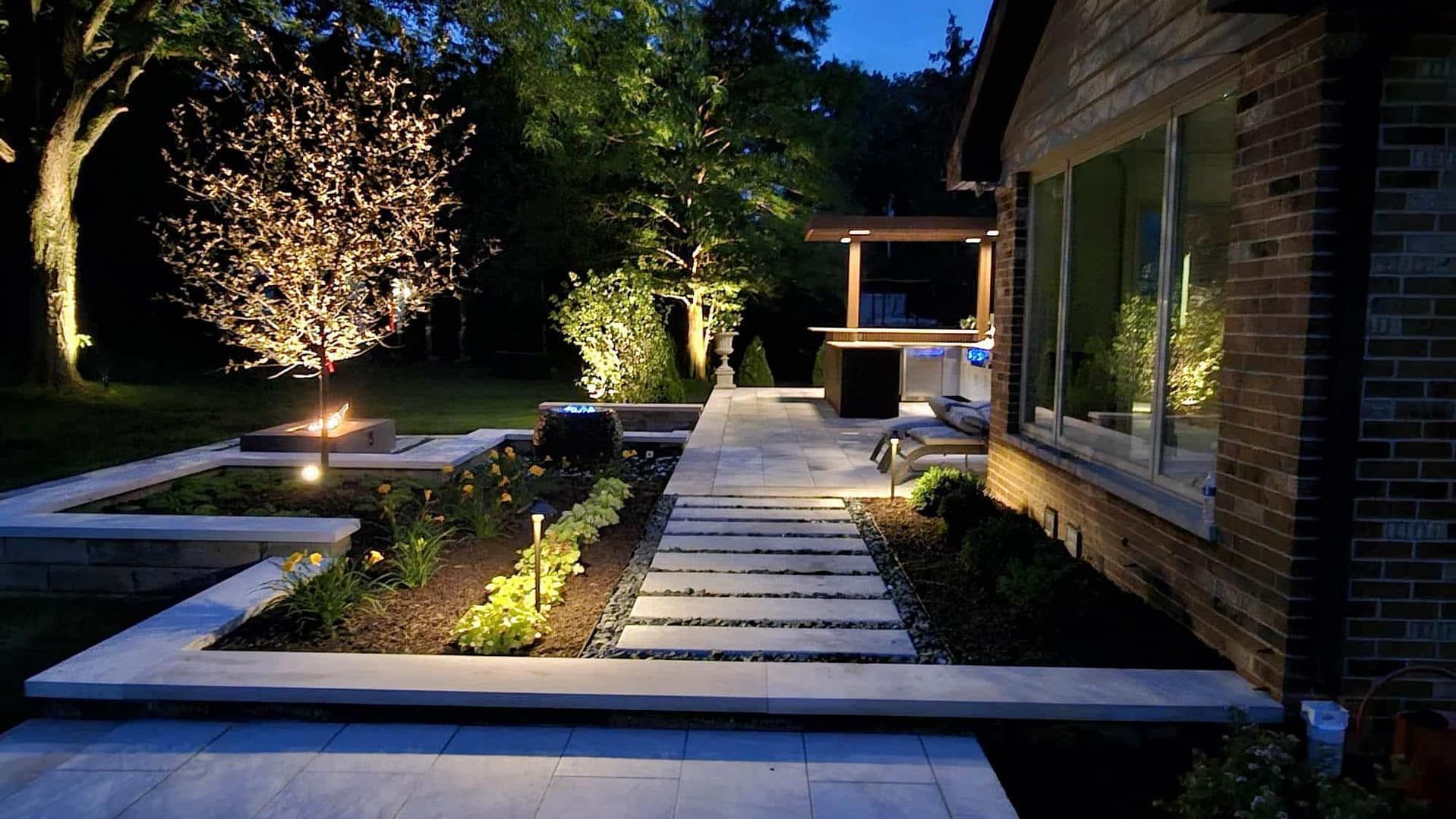 Nighttime view of illuminated backyard patio with fire pit, garden, and house.