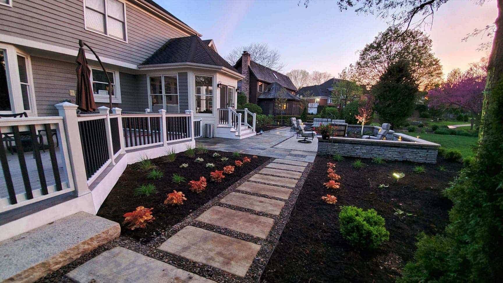 Backyard with a stone path, deck, landscaping, and a fire pit at sunset.