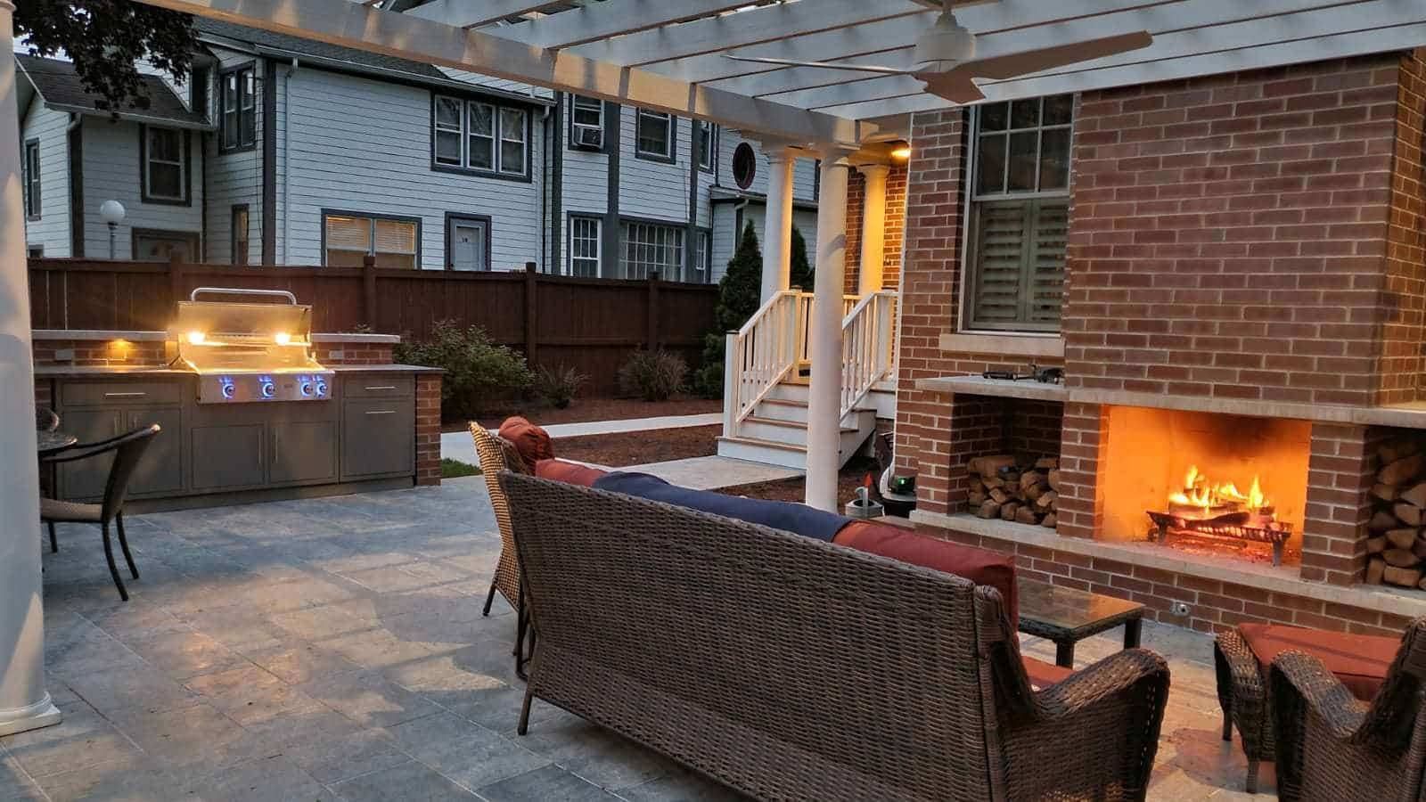 Outdoor patio with a fireplace and grill at dusk. Wicker furniture, brick, and a pergola are visible.
