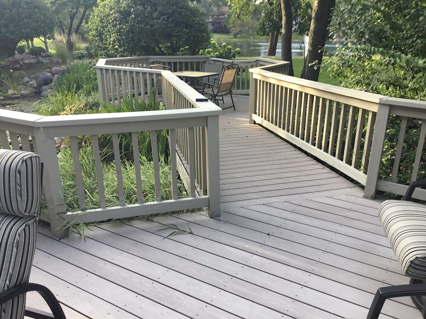Wooden deck with railing, table, and chairs overlooking a pond and lush greenery.