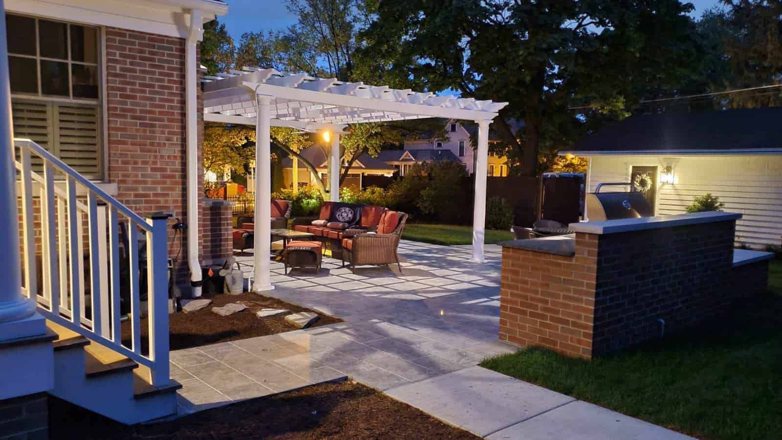 Backyard patio with a white pergola, seating, and brick grill area at dusk.