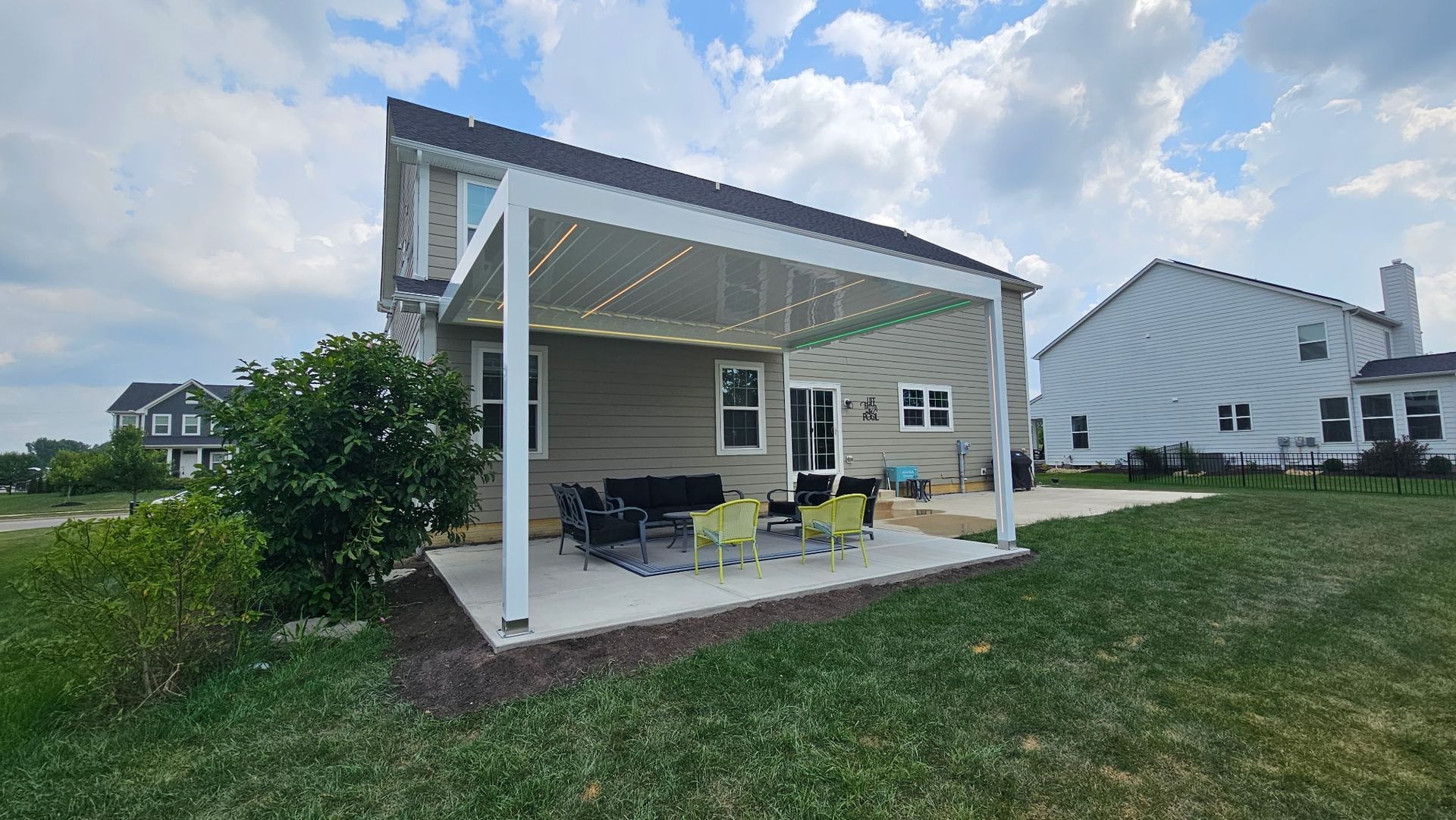 Backyard patio with white pergola, furniture, and house under a cloudy sky.
