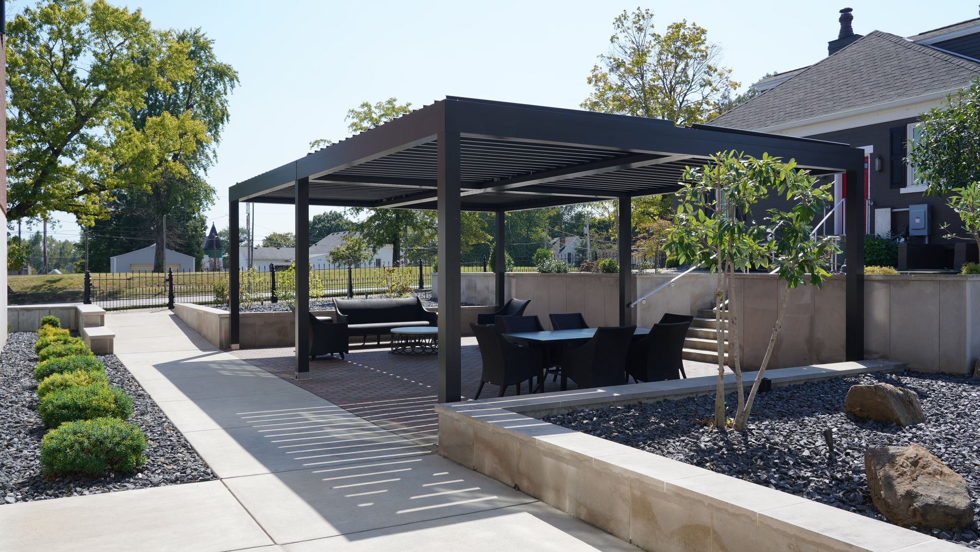 Patio with black pergola, tables, and seating. Gravel ground, stone planters with shrubs.