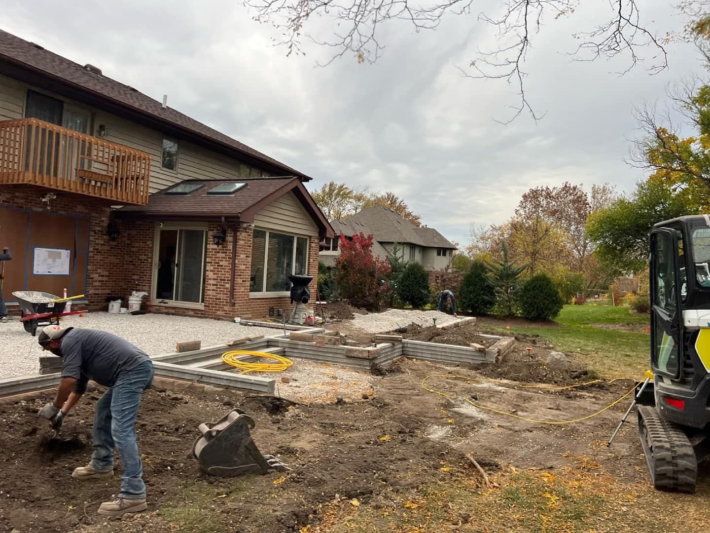 Construction worker shovels dirt near a house with a patio being built. A small excavator is on the right.
