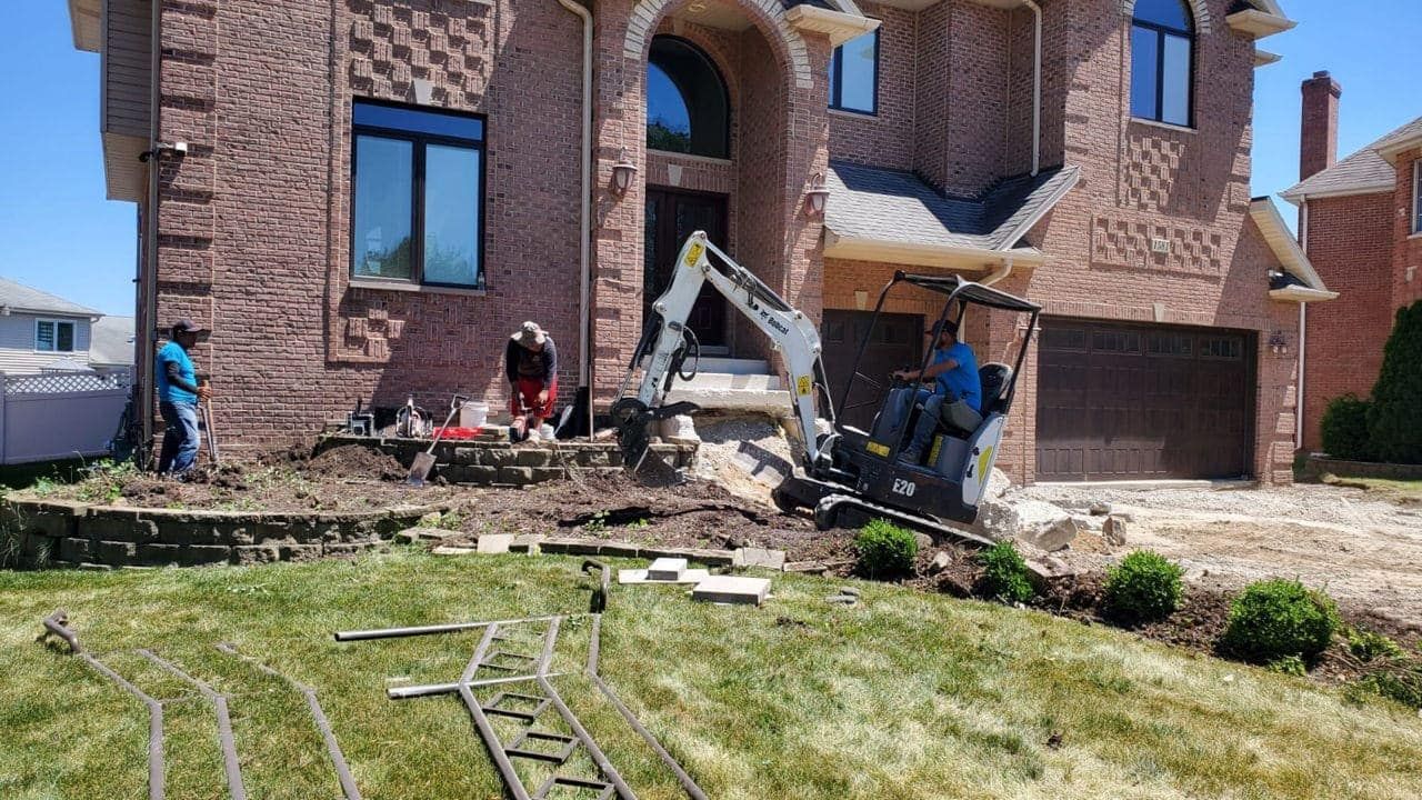 Construction workers landscaping a brick house with an excavator on a sunny day.