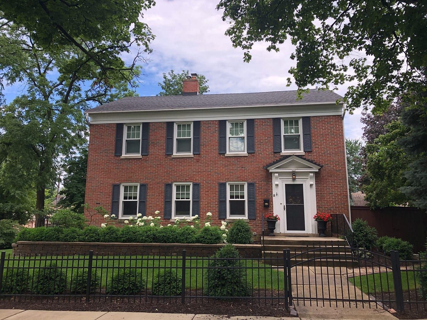 Red brick two-story house with black shutters, a black door, and a small front yard with a black fence.