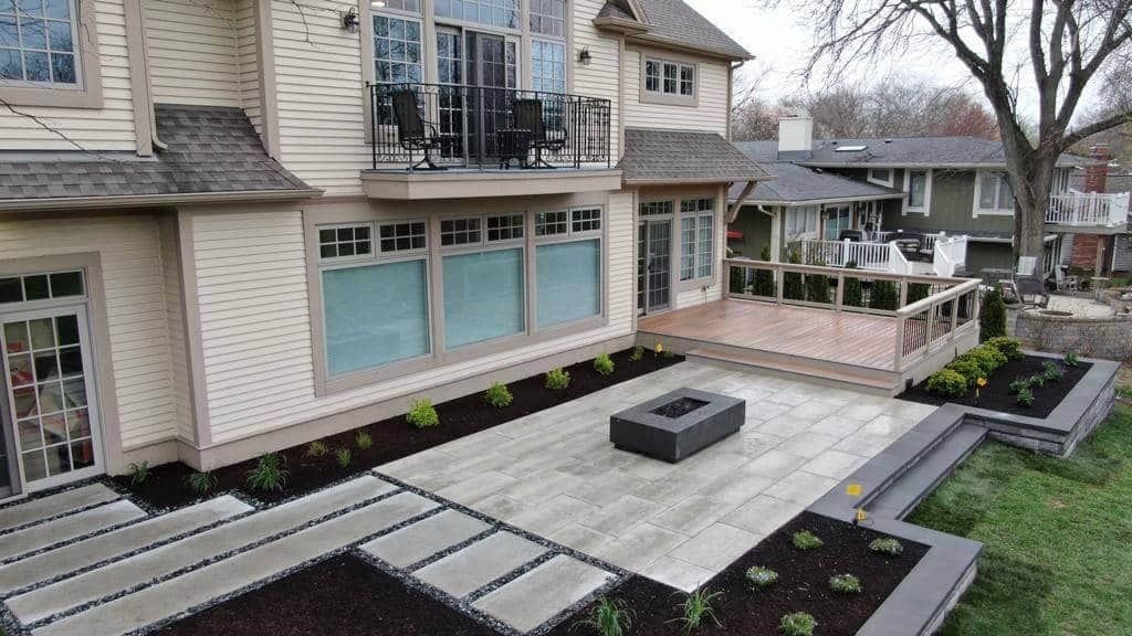 Backyard patio with fire pit, deck, and house. Beige siding, stone pavers, and lush landscaping.