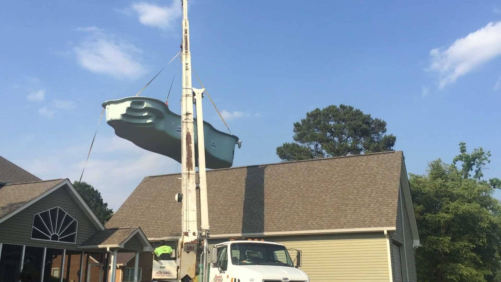A truck crane lifting a prefabricated swimming pool over a house with a brown roof.