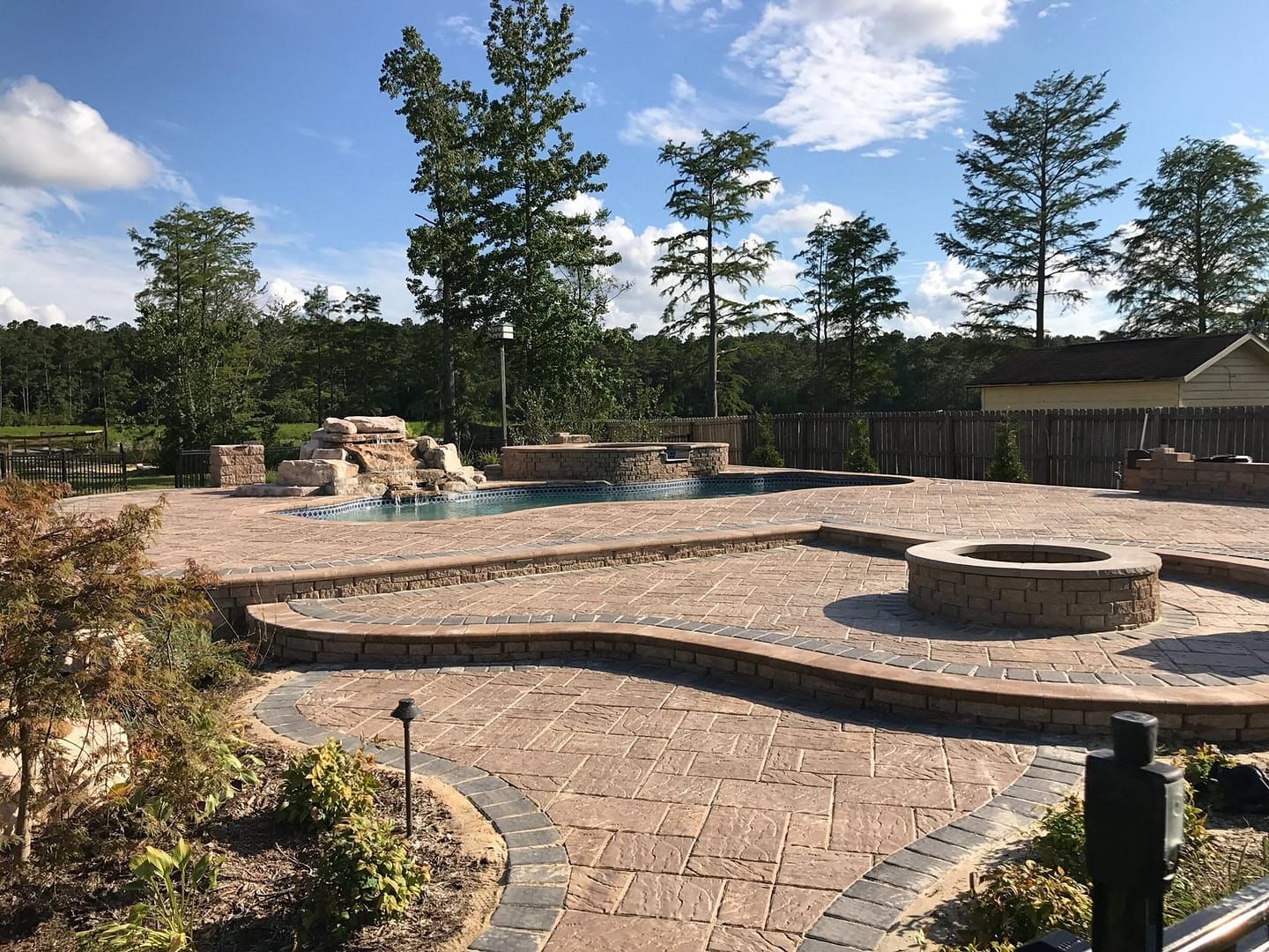 Stone patio with pool, fire pit, and trees under a blue sky.