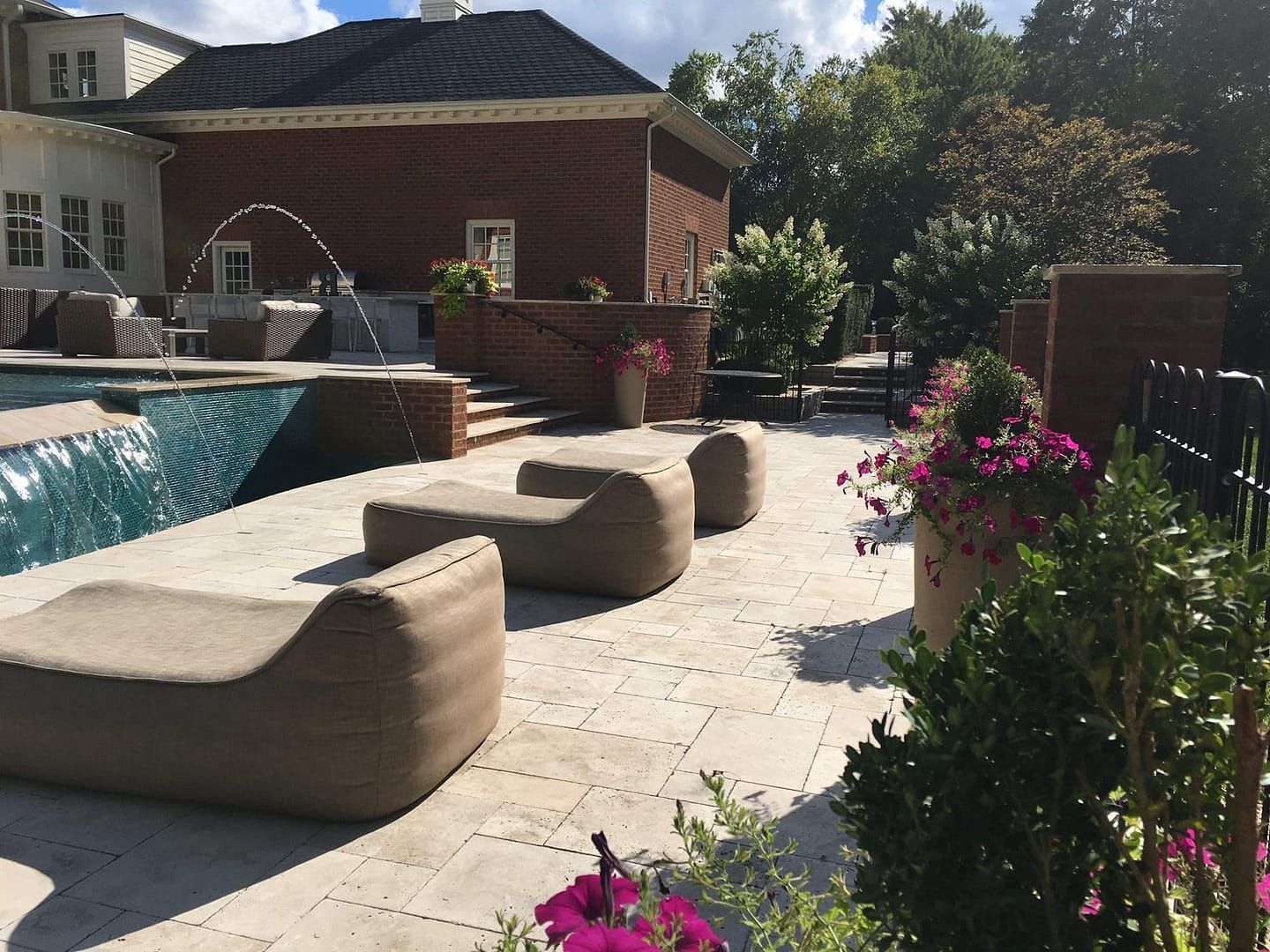 Patio with pool, lounge chairs, brick wall, and pink flowers under a sunny sky.