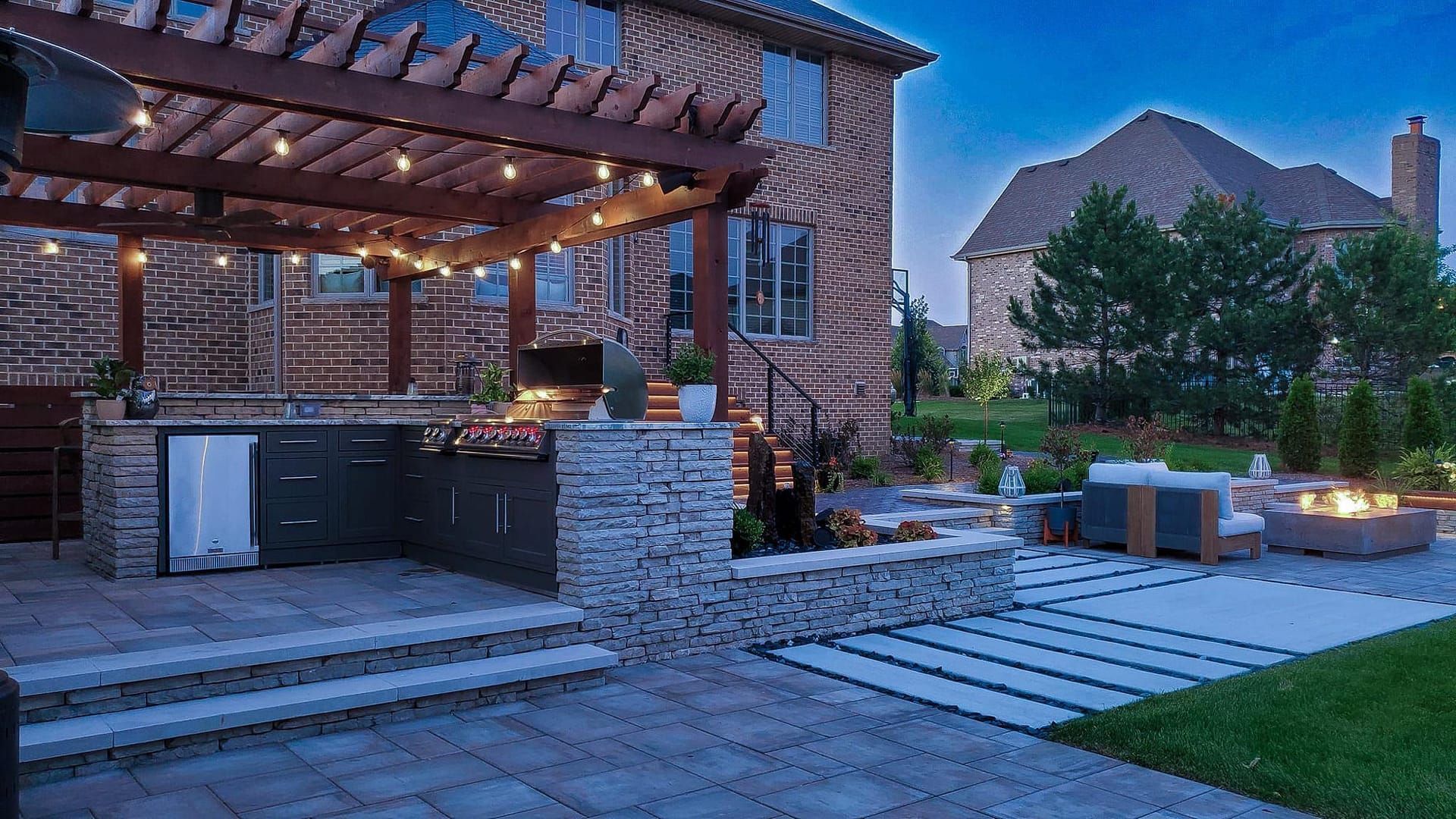 Outdoor kitchen with pergola, lights, and seating area by a brick house at dusk.