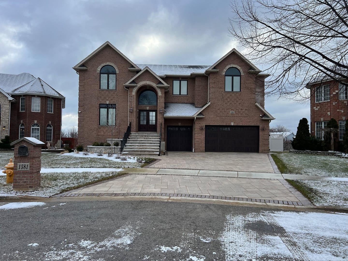 Brick house with two-car garage and snow-covered lawn under a cloudy sky.