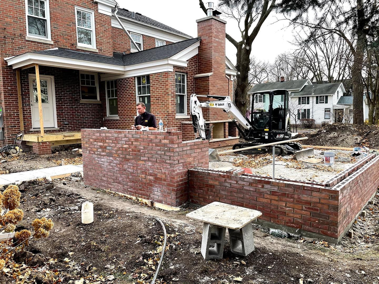 Brickwork construction in progress next to a house. A person stands near a small excavator.