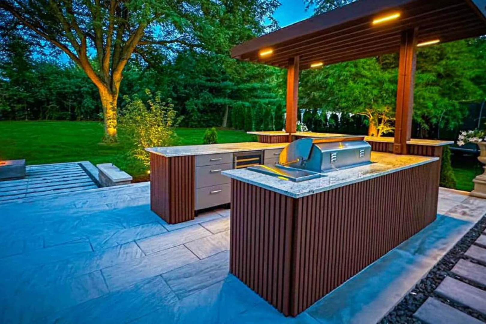 Outdoor kitchen with grill under a wooden pergola, on a stone patio, surrounded by a lawn and trees.