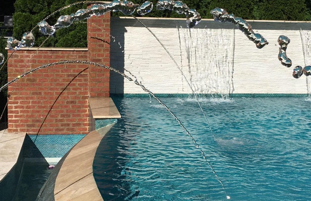 Pool with arched water jets, brick and white stone wall backdrop.