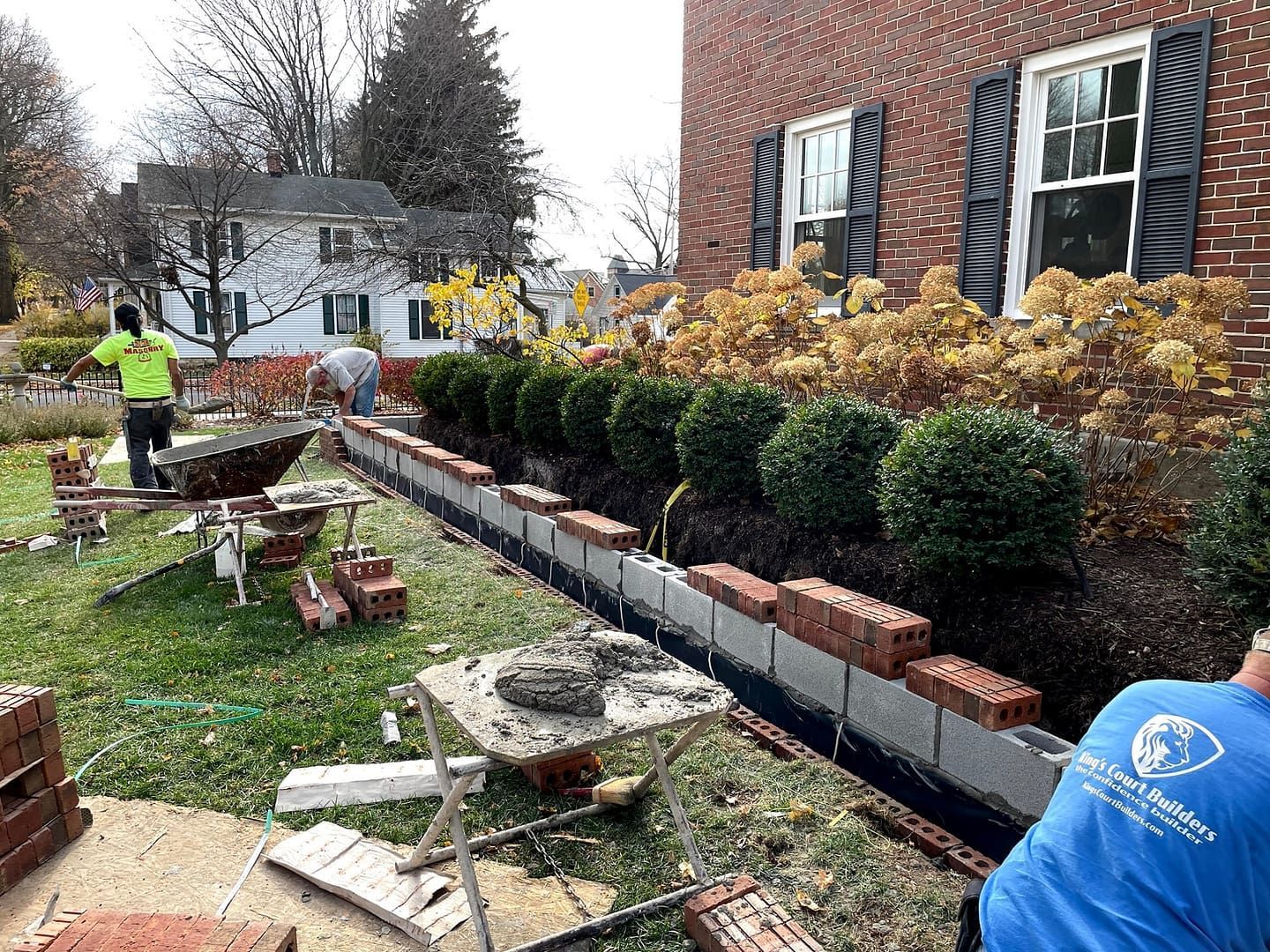 Construction workers building a brick border in front of a brick building. Green lawn and shrubs.