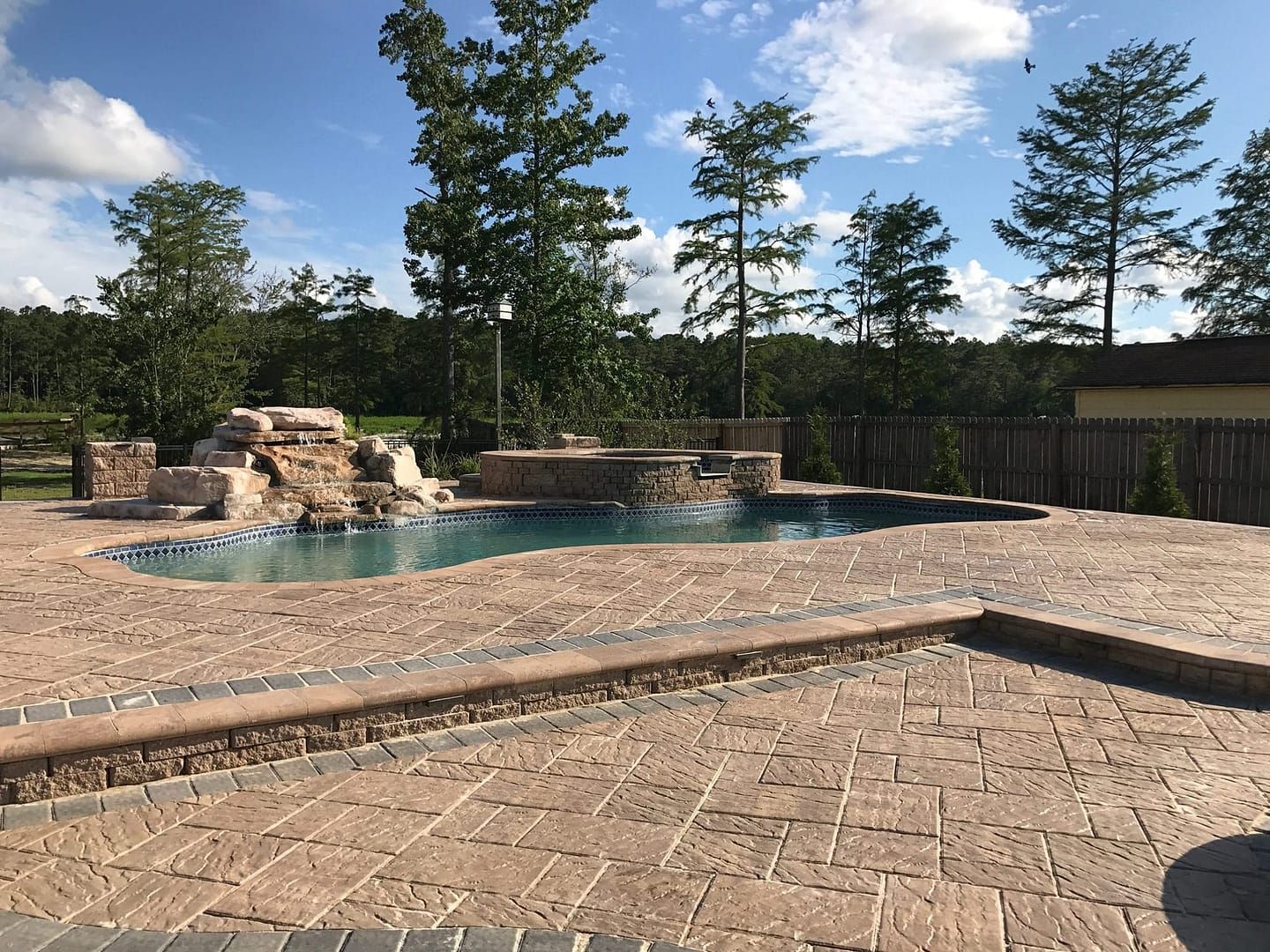 A backyard with a pool and a brick patio, with trees and a cloudy sky.