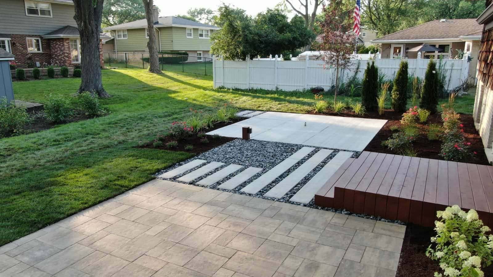 Backyard patio with pavers, stepping stones, and a wooden deck, surrounded by greenery and flowers.