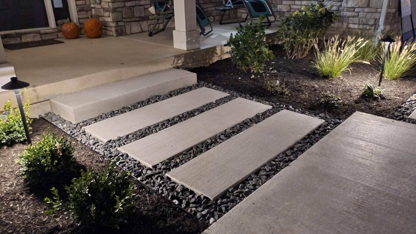Concrete walkway with stepping stones, bordered by dark rocks and lit by low-voltage lights.