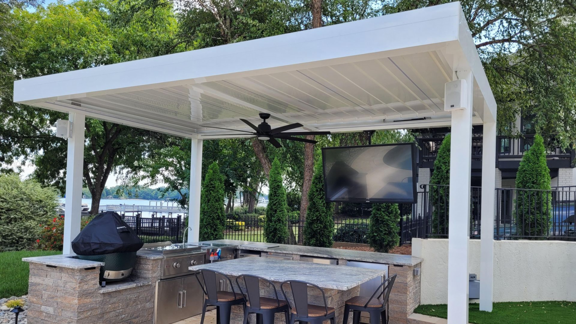 White outdoor kitchen with a grill, bar, TV, and overhead fan. Lakeside setting.
