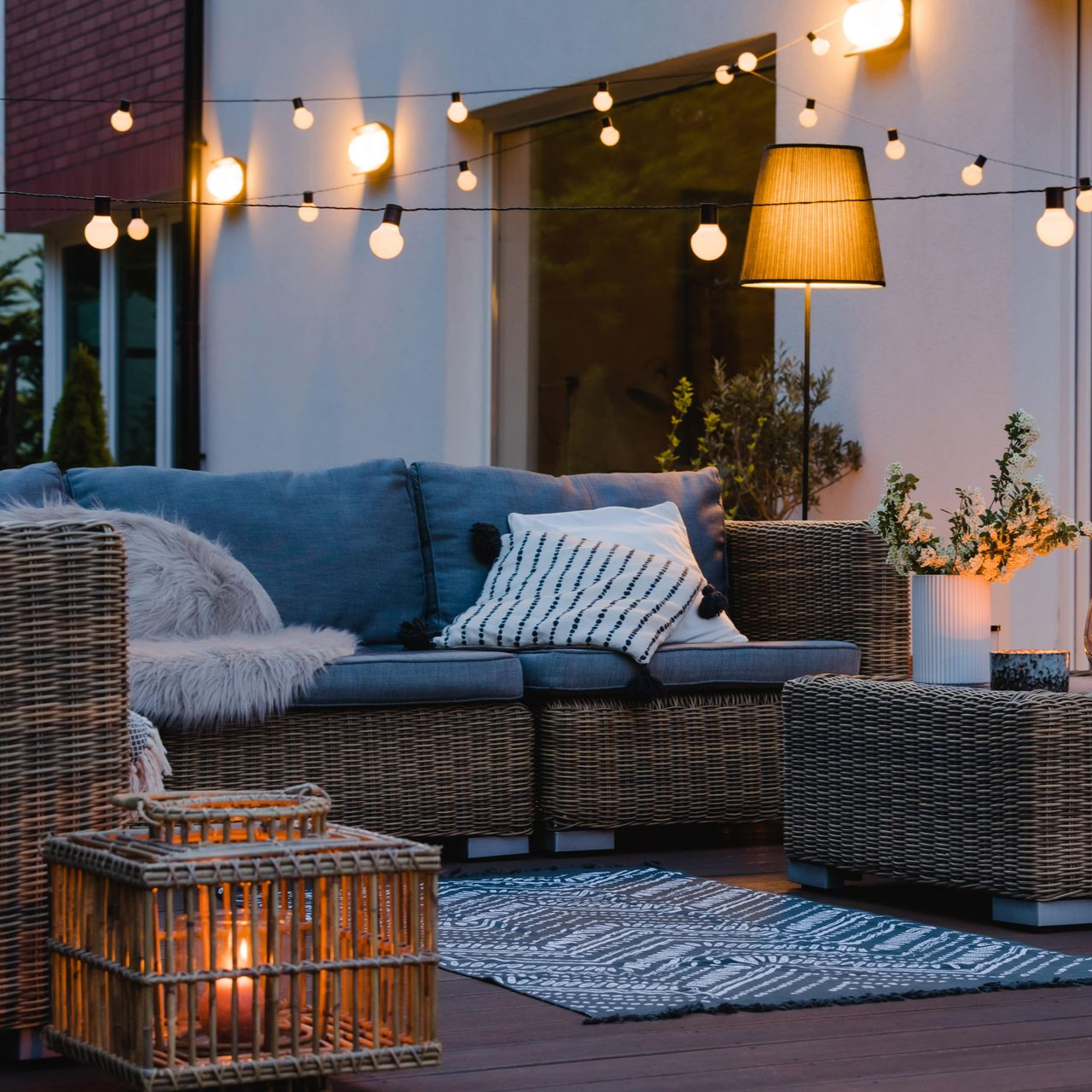 Outdoor patio with string lights, wicker furniture, cushions, rug, and lantern.