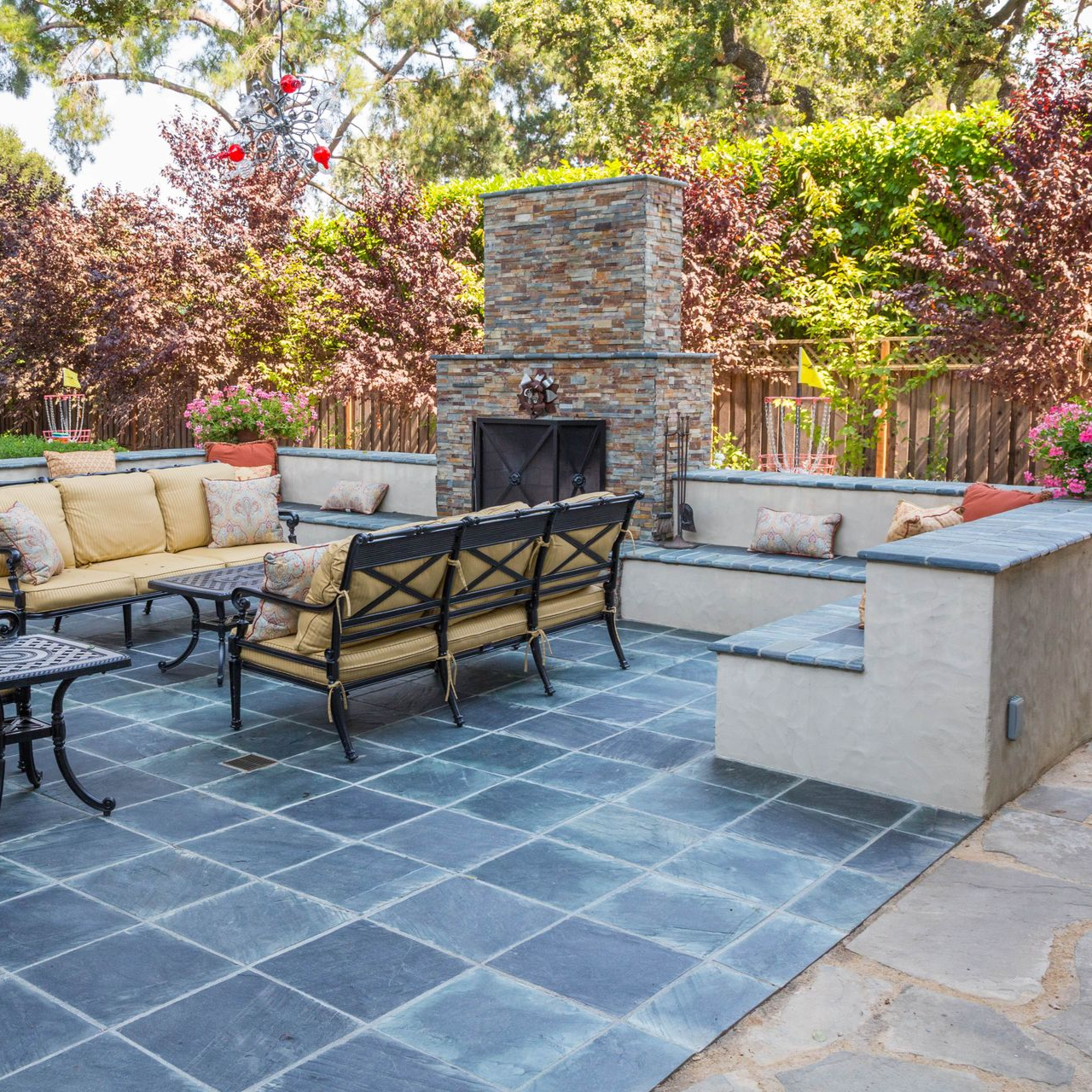 Patio with couches, fireplace, built-in seating, blue tile flooring, surrounded by lush greenery and trees.