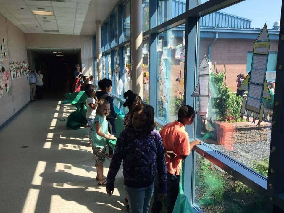 Children looking out a large window, presumably at a garden, from a school hallway.