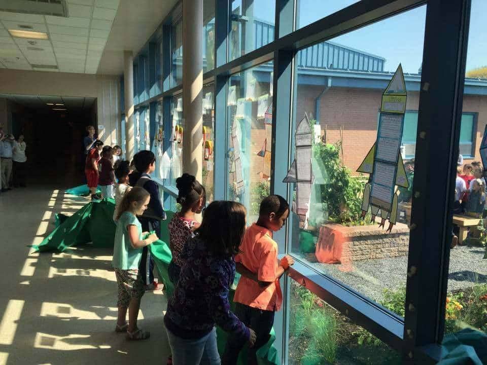 Children looking out a large window at a garden; some holding green bags, others looking at charts.