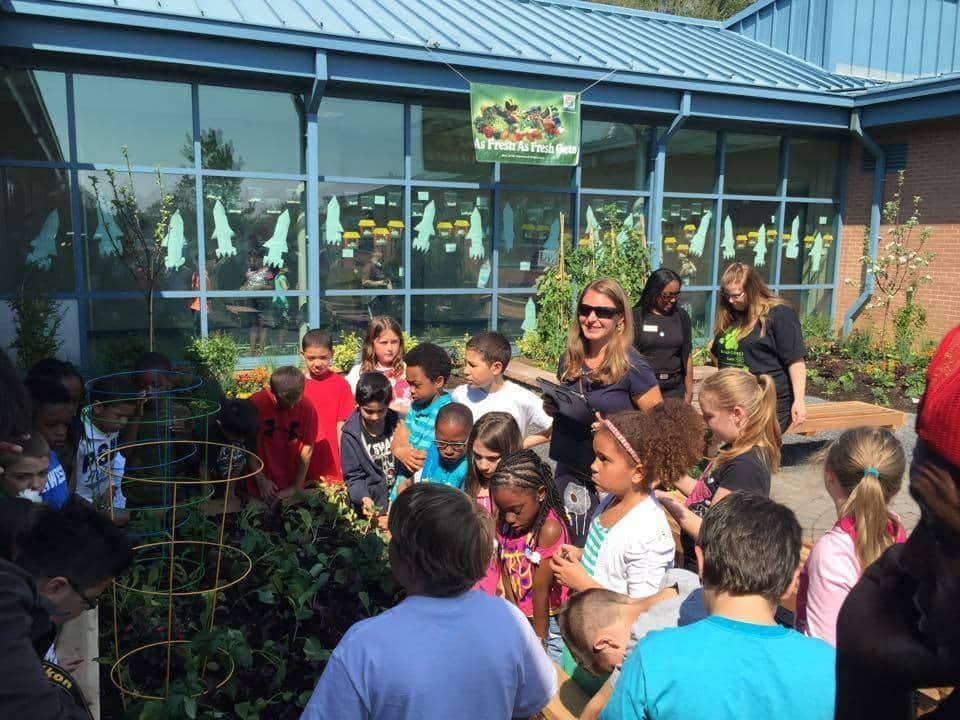 Children and adults tending a school garden; blue building, sunny day.