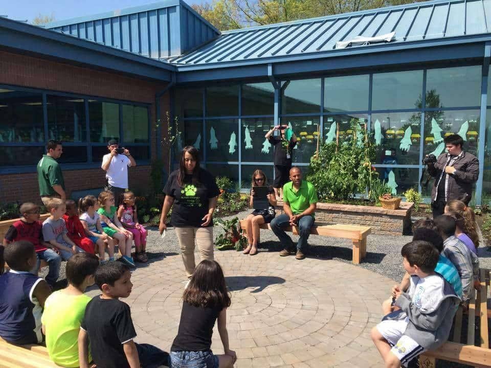 Children listen to a presentation in an outdoor courtyard, with adults and plants visible.