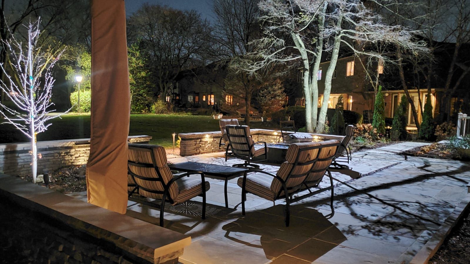 Patio scene at night, snow-covered chairs around a fire pit. Light illuminates the area and trees.