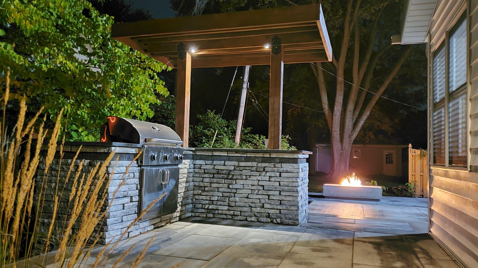 Backyard with outdoor kitchen, fire pit, and wooden pergola lit at night.