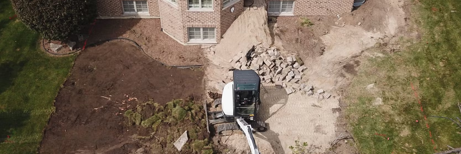An aerial view shows construction work near a brick house. A small excavator is in the middle of a dirt yard.