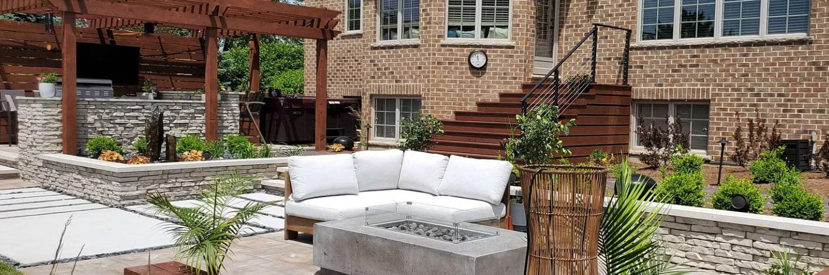 Backyard patio with a sectional sofa, fire pit, and outdoor kitchen under a pergola. Brick house and steps visible.