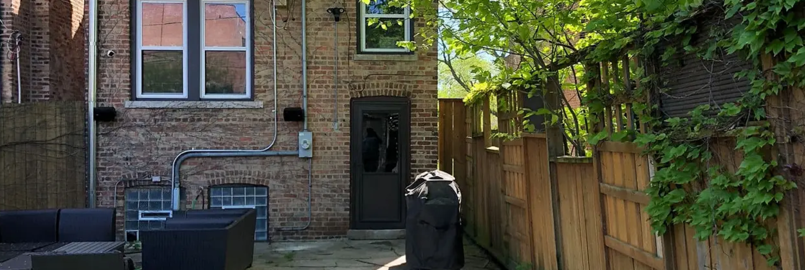 Brick building backyard with a wooden fence covered in green vines. There's a grill and patio furniture.