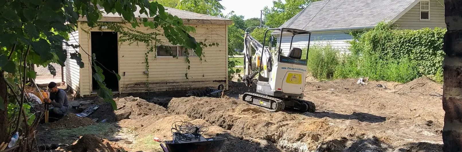 An excavator digging a trench in front of a small building, with a second building in the background.