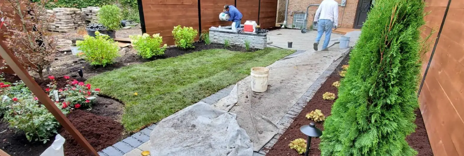 Landscapers working on a backyard. Green grass, plants, and a stone pathway. Wooden fence and walls.