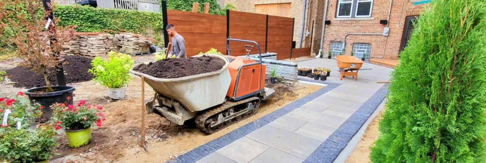 A small tracked dumper transporting soil in a backyard under construction. Plants and a patio are in view.